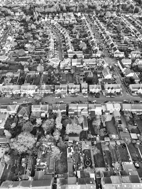 An aerial black-and-white photograph of a residential area featuring a variety of gardens, lawns, and pathways among rows of terraced and semi-detached houses. In the foreground, there are back garden