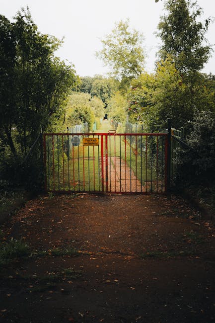 A red metal gate blocks the entrance to a garden pathway on a residential estate, with a small sign attached indicating restricted access. Beyond the gate, the pathway continues straight, bordered by